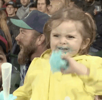 a little girl with pig tails in a yellow rain coat at a baseball game eating blue cotton candy and exhibiting the most excited face you can imagine